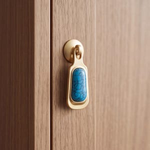 Puede incluir: Un tirador de metal en tono latón con una placa de montaje redonda y lisa y un colgante vertical rectangular. El colgante está incrustado con una piedra lisa de forma ovalada en color turquesa que presenta un patrón oscuro de vetas.