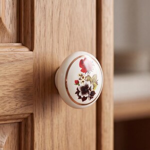 May include: A round, white ceramic cabinet knob featuring a central floral illustration with red, dark purple, and yellow flowers and green leaves. The knob is accented with a thin, metallic copper-coloured circular rim and is mounted on a wooden door.