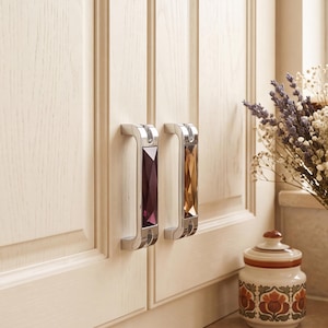 May include: Two rectangular cabinet pulls with a polished silver-tone metal frame and a faceted, jewel-like center insert. One pull features a deep purple insert, while the other features a warm amber-colored insert, both mounted on light wood doors.
