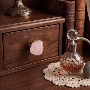 May include: A drawer knob featuring a raw, pale pink rose quartz stone with a metallic gold-toned edge, mounted on a dark brown wooden drawer. Beside it sits a clear glass perfume bottle with a faceted stopper and a gray fabric bulb pump.