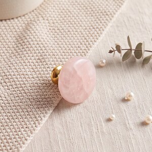 May include: A round cabinet knob featuring a polished, translucent pale pink stone face with a smooth, rounded surface. The knob is attached to a circular, shiny gold-toned metal base that connects to the back of the stone, providing a sturdy mount.