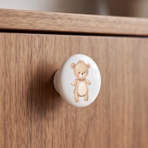 Pode incluir: Um puxador de móvel redondo em cerâmica branca com uma ilustração central de um urso de peluche castanho-claro com bochechas cor-de-rosa. O urso está representado de pé com uma mancha bege mais clara na barriga, orelhas e nariz escuro.