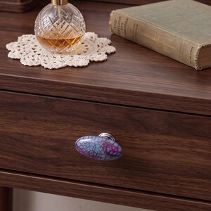 May include: An oval-shaped drawer knob featuring a crackled pattern in shades of purple and turquoise with a silver-toned metal base, attached to a dark brown wood-grain drawer front with a horizontal wood texture and a smooth finish.