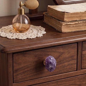 May include: A dark brown wooden drawer featuring a single cabinet knob made from a raw, purple amethyst crystal attached to a gold-toned metal base. The drawer is part of a wooden furniture piece with a visible wood grain texture and finish.