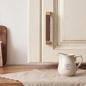 May include: A cabinet handle featuring a dark brown wood center section with brushed gold metal caps at the top and bottom. The handle is mounted vertically on a cream-colored cabinet door with a visible wood grain texture and recessed paneling.