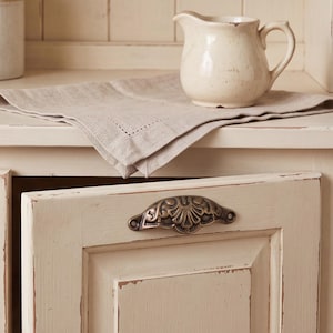 May include: A cream-coloured ceramic creamer pitcher sits on a folded light beige linen napkin. The pitcher is placed on a cream-coloured cabinet door featuring a decorative, ornate, antique-style bronze metal cup pull handle with a shell motif.