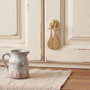 May include: A matte gold metal drop pull handle with a vertical, elongated teardrop shape mounted on a cream-colored cabinet door with a distressed finish, positioned next to a small, rustic, white and brown ceramic pitcher on a linen cloth.