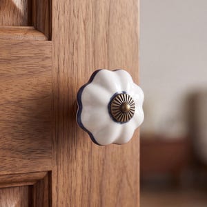 May include: A white ceramic cabinet knob with a scalloped, flower-like shape and a dark blue rim. The center of the knob features a circular, metallic bronze-toned decorative plate with a radial, ribbed pattern, attached to a wooden door.