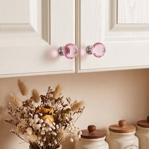 May include: Two round, faceted pink glass cabinet knobs with polished silver-tone metal bases. The knobs are attached to white cabinet doors, featuring a textured, multi-faceted surface that reflects light, creating a clear, jewel-like appearance.
