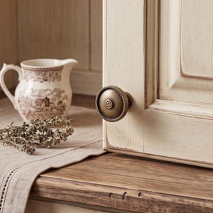 Peut inclure: Un bouton de placard rond en laiton antique avec un bord cannelé et un petit détail circulaire central présentant un motif floral. Le bouton est fixé sur une porte de placard en bois crème vieilli avec une texture de grain de bois visible.