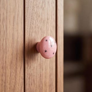 May include: A round, glossy, light pink ceramic cabinet knob featuring a pattern of small, dark, four-pointed star shapes. The knob is attached to a vertical wooden panel with a visible grain texture, showing a light brown natural wood finish.