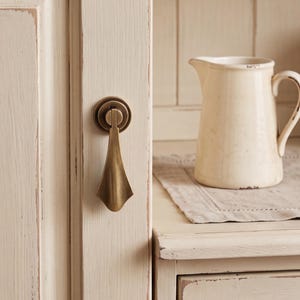 May include: A brass-toned metal drop pull handle mounted on a cream-colored wooden cabinet door. Beside the cabinet, a cream-colored ceramic pitcher sits on a light-colored linen cloth, completing the scene with a neutral color palette.