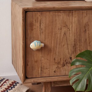 May include: A ceramic cabinet knob shaped like a small fish with a pale yellow body and light blue stripes. The fish-shaped knob is mounted on a wooden cabinet door with a visible wood grain texture, featuring a single dark eye detail.