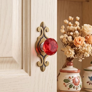 Puede incluir: Un pomo de armario de cristal rojo facetado montado sobre una placa metálica ornamentada de color latón antiguo con detalles de volutas. El pomo está fijado a una puerta de armario de madera clara, con un acabado pulido y una forma geométrica.