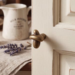 May include: A brass-toned metal cabinet knob with an organic, cross-like shape featuring rounded, smooth edges. The knob is mounted onto a cream-colored wooden cabinet door with a visible grain and distressed edges, showing a darker base layer.