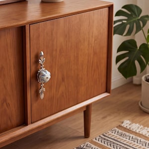 May include: A wooden cabinet featuring a vertical metal handle with a decorative, round, white ceramic knob. The knob is adorned with a blue and green floral pattern. The cabinet has a smooth, light brown wood grain finish and stands on legs.