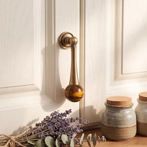 May include: A brass cabinet pull featuring a vertical tapered handle attached to a circular base, finished with a spherical tiger's eye gemstone at the bottom. The hardware is mounted on a white paneled cabinet door with a smooth metallic sheen.