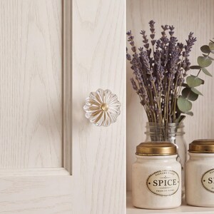 May include: A clear, fluted glass cabinet knob with a central brass-toned metal accent, mounted on a light-colored wood cabinet door. Beside it, a glass jar holds dried lavender, and two cream-colored ceramic spice jars sit on a shelf.