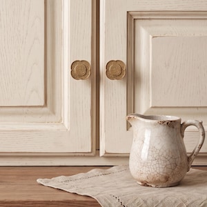 Puede incluir: Dos pomos de armario color latón con forma floral de cuatro lóbulos y detalles en relieve, montados en puertas de armario de madera color blanco roto, acompañados por una pequeña jarra de cerámica color crema con esmalte craquelado.