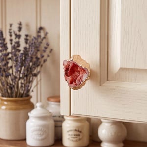May include: A cabinet knob featuring a raw, irregular red geode stone center with a textured, metallic gold-toned edge. The stone displays a crystalline, deep red interior texture, mounted onto a light-colored, paneled wooden cabinet door.