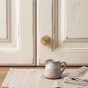 May include: A small, round, gold-colored metal cabinet knob with a sunburst or flower-like radial pattern. The knob is attached to a cream-colored cabinet door with a distressed finish. A small, two-toned ceramic creamer sits on a linen cloth.