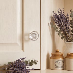 May include: A clear, faceted, octagonal glass cabinet knob mounted on a light cream-colored wooden cabinet door. The knob features a central starburst pattern and is positioned next to dried lavender sprigs and two small ceramic storage jars.