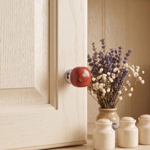 May include: A round, polished, reddish-brown stone cabinet knob with a silver-toned metal base, mounted on a light-colored, wood-grained cabinet door. The knob features natural marbling and a smooth, glossy finish, providing a distinct contrast.