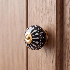 以下が含まれることがあります： 光沢のある黒い表面に白い放射状の花柄が施され、中央にギザギザの縁取りがある金色の金属製ディスクが付いた丸いキャビネットノブ。ノブは、縦の木目が目立つ明るい茶色の木製表面に取り付けられています。