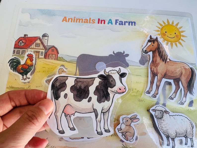 Interactive farm animal activity page showing a child's hand holding a cow cutout ready to be matched on the laminated barnyard scene.