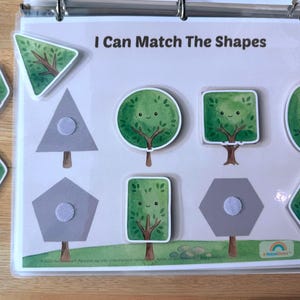 May include: A learning activity sheet titled I Can Match The Shapes featuring a light background with gray shape outlines and brown tree trunks. Several removable tree-shaped pieces with green foliage and smiling faces are included for matching.