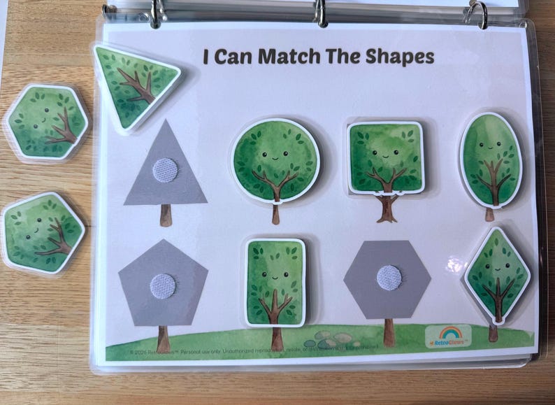 May include: A learning activity sheet titled I Can Match The Shapes featuring a light background with gray shape outlines and brown tree trunks. Several removable tree-shaped pieces with green foliage and smiling faces are included for matching.