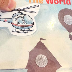 Close-up of "The World of Vehicles" activity page showing a helicopter cutout being placed on a sky-themed background. Rough hook velcro dots were applied to the book and soft loop dots to the cutout piece.