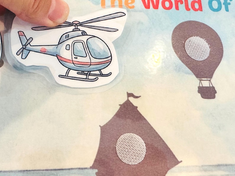 Close-up of "The World of Vehicles" activity page showing a helicopter cutout being placed on a sky-themed background. Rough hook velcro dots were applied to the book and soft loop dots to the cutout piece.