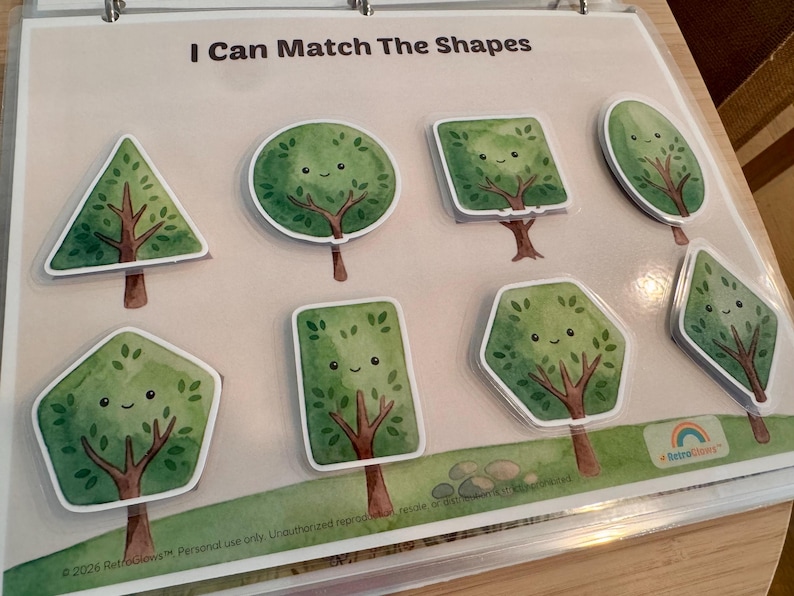 May include: A laminated educational activity sheet titled I Can Match The Shapes featuring seven green tree illustrations in various geometric forms including a triangle, circle, square, oval, pentagon, rectangle, hexagon, and diamond, each with a smiling face.