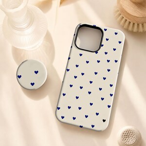 Pode incluir: Uma capa de smartphone na cor creme e um suporte circular para telemóvel a condizer, ambos com um padrão repetitivo de pequenos corações azul-escuros. A capa tem um recorte retangular grande para a lente da câmara e um rebordo preto.