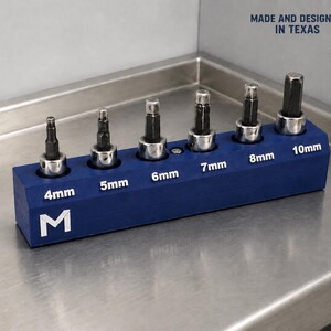 May include: A blue rectangular tool organizer holding six metal hex bits in ascending sizes from 4mm to 10mm. The base features white text labels for each bit size, a white letter M logo, and the text MADE AND DESIGNED IN TEXAS with a Texas flag.