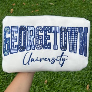 May include: A white sweatshirt featuring the word GEORGETOWN in large, block-style letters filled with various blue and white patterns, including stripes, polka dots, and florals. Below, the word University is embroidered in a blue cursive script.