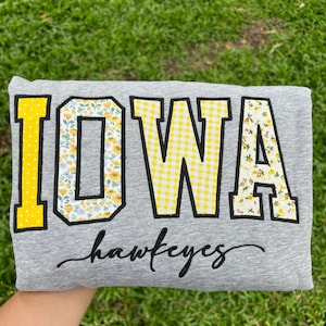 May include: A heather gray sweatshirt featuring the word IOWA in large block letters with black outlines. The letters I, O, and A have a yellow and white floral pattern, while the letter W has a yellow and white gingham pattern. The word hawkeyes is embroidered in black cursive script below.
