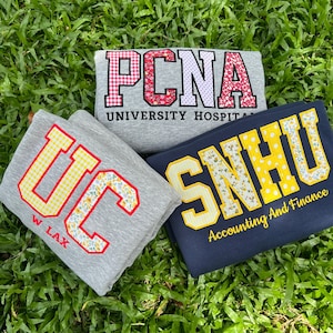 May include: Three folded sweatshirts featuring collegiate-style applique lettering. One gray shirt displays PCNA UNIVERSITY HOSPITAL, one gray shirt shows UC W LAX, and one navy blue shirt features SNHU Accounting And Finance with patterned fabric.