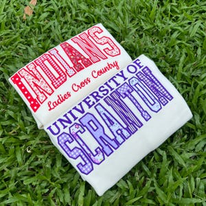 May include: Two white t-shirts with graphic prints. One shirt features the red text INDIANS and Ladies Cross Country. The other shirt features the purple text UNIVERSITY OF SCRANTON with a checkered and floral pattern inside the block letters.