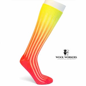 Può includere: Un calzino al ginocchio con una trama a coste verticali e una sfumatura di colori vivaci che passa dal giallo brillante del bordo superiore all'arancione e al rosso intenso della punta. Il logo WOOL WORKERS con due icone di pecore è visibile.
