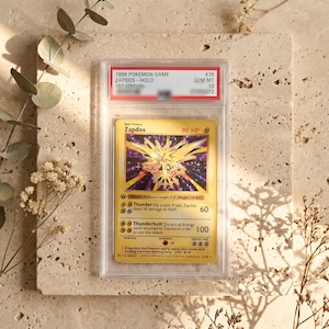 May include: A 1999 Pokemon Game Zapdos holographic trading card, graded GEM MT 10, encased in a clear protective plastic slab. The card features the yellow Zapdos character, 90 HP, and the attacks Thunder and Thunderbolt with text details.