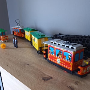 May include: A collection of toy trolley cars featuring red and yellow bodies with green roofs, numbered 3, accompanied by two small figurines, a yellow circular accessory, and a section of black plastic train tracks arranged in a line.