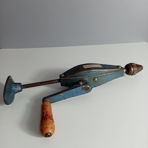 Pode incluir: Um berbequim manual vintage com corpo de metal azul texturizado com ferrugem visível, um cabo de madeira com acabamento gasto e uma bucha de metal na frente. A ferramenta inclui uma placa de peito de metal e um mecanismo de manivela.