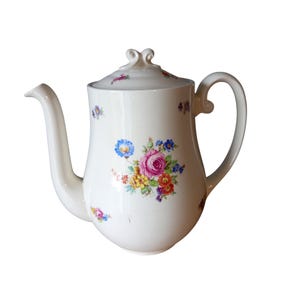 May include: A white porcelain teapot featuring a floral pattern with pink, yellow, blue, and orange flowers. The teapot has a curved handle, a long spout, and a lid with a decorative finial. The surface is decorated with small floral sprigs.