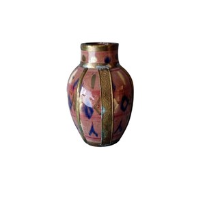 Może przedstawiać: Mały ceramiczny wazon w kolorze przygaszonego różu, z pionowymi złotymi metalowymi pasami i ozdobnym złotym brzegiem. Powierzchnia ozdobiona jest powtarzającym się wzorem małych niebieskich rombów oraz rozproszonymi złotymi pociągnięciami pędzla.