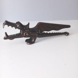 May include: A dark bronze-colored metal nutcracker shaped like a dragon with an open mouth, textured wings, and a long, serrated tail. The dragon's head forms the lever mechanism, while the body and tail extend horizontally to form the handle.