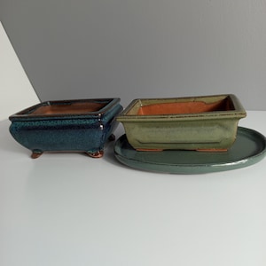 Puede incluir: Dos macetas rectangulares de cerámica para bonsái. Una maceta presenta un esmalte azul profundo y brillante con cuatro patas pequeñas. La segunda tiene un esmalte verde mate y se asienta sobre un plato ovalado a juego. Ambas tienen interior de terracota.