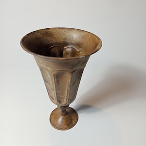 Può includere: Un calice in metallo color bronzo usurato con un bordo ampio e svasato e un corpo scanalato e sfaccettato che si restringe verso uno stelo sottile e una base circolare. La superficie presenta una patina invecchiata e maculata.