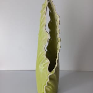 Può includere: Un vaso alto in ceramica color verde chiaro con una struttura verticale simile a una foglia. L'oggetto presenta una finitura liscia e lucida e un bordo ondulato e arricciato distintivo che corre lungo l'apertura, creando una forma organica allungata.